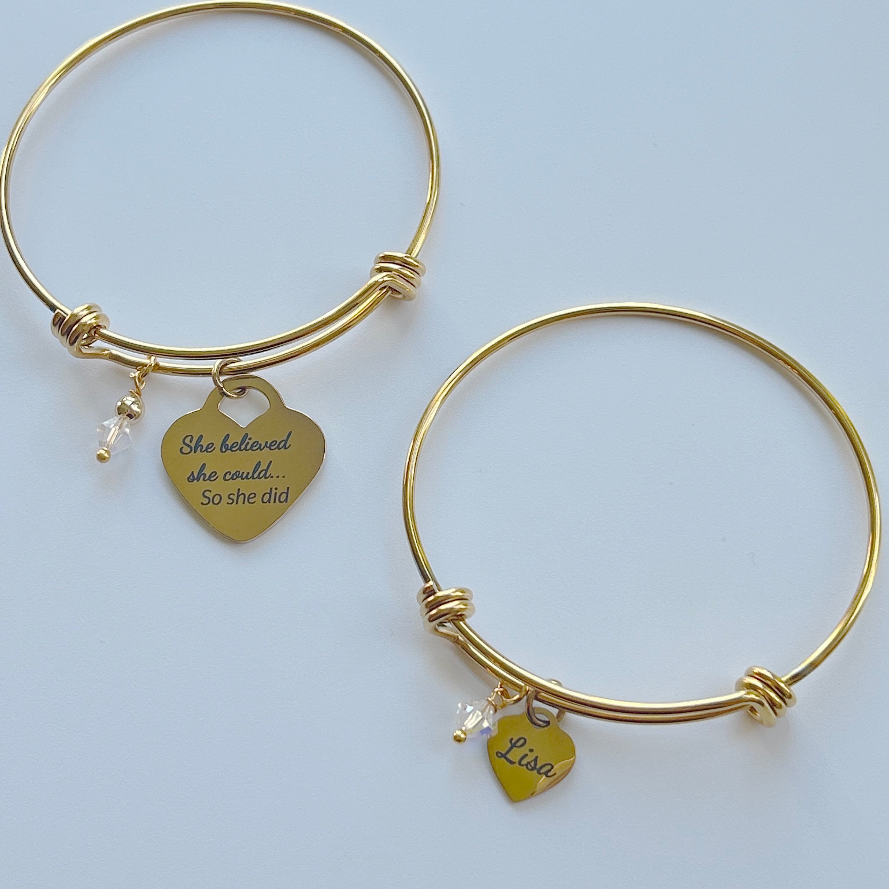 this image shows two bangle style bracelets , both with a clear crystal bead accent; one features a Large heart charm saying "She believed she could so she did", the other has the name "Lisa"