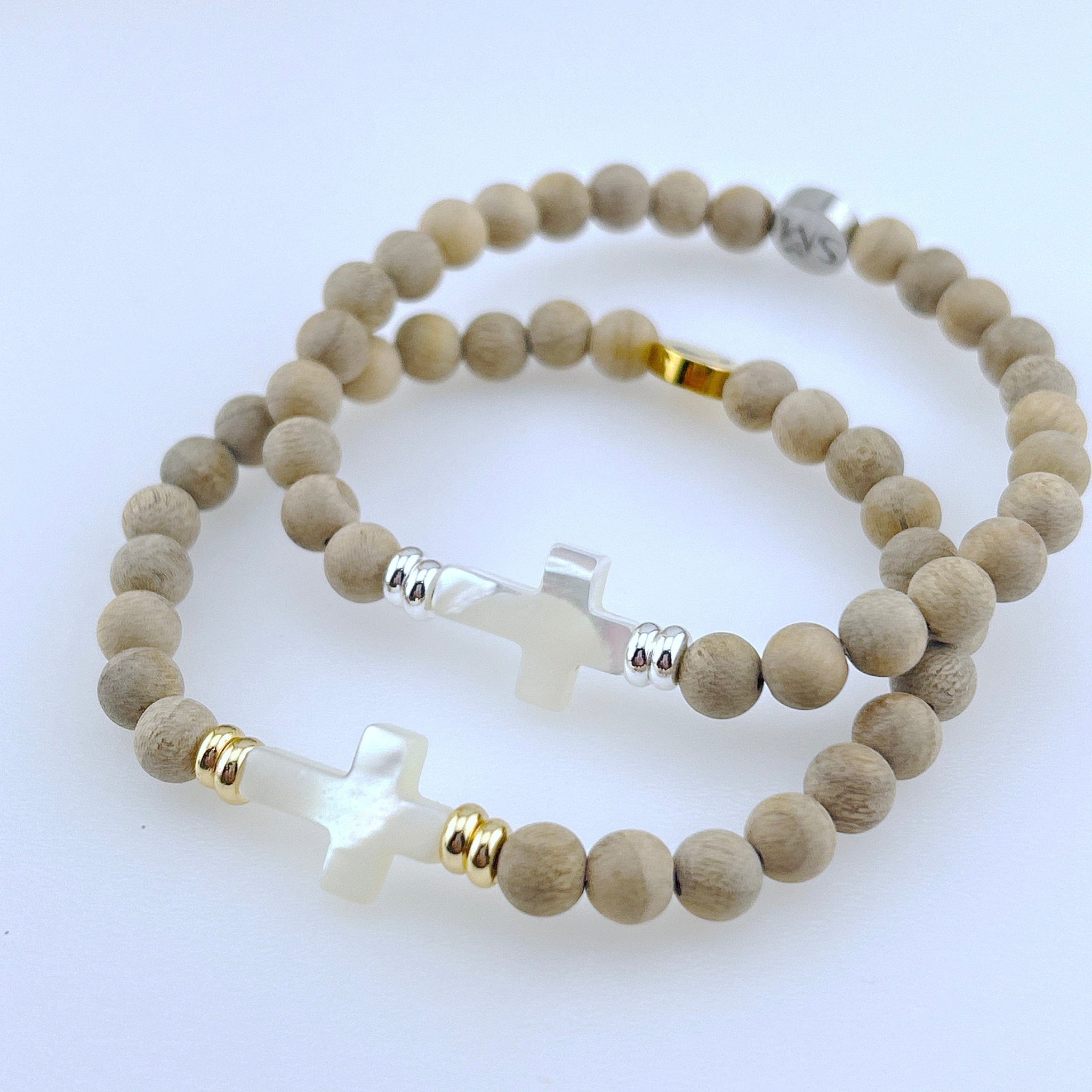white background, 2 wooden bead bracelts stacked on top of each other, both with a mother of pearl cross, and one bracelet with gold bead accents and the other with silver bead accents.
